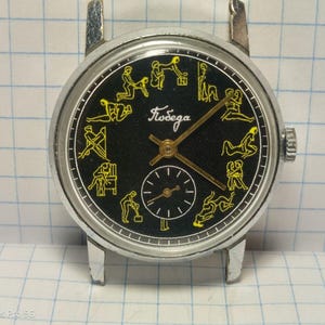 May include: A vintage wristwatch with a black face and silver-tone case. The watch face features yellow illustrations of various figures around the perimeter and the word "Победа" in cursive. A smaller dial is at the bottom.