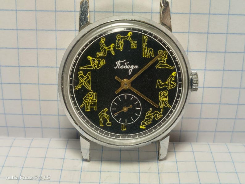 May include: A vintage wristwatch with a black face and silver-tone case. The watch face features yellow illustrations of various figures around the perimeter and the word "Победа" in cursive. A smaller dial is at the bottom.
