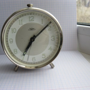 May include: A silver alarm clock with a white face and black hands. The clock has a silver trim and a black knob on the side. The clock face has the word "PRIM" printed on it.
