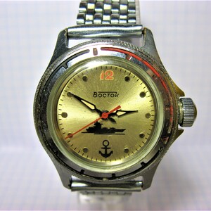 Puede incluir: Un reloj de pulsera plateado con una esfera dorada y agujas negras. El reloj tiene un segundero rojo y un logotipo negro de ancla y barco. La esfera del reloj dice "Vostok" y "12".