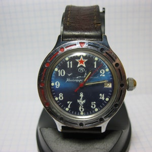 Puede incluir: Un reloj de pulsera soviético vintage con una esfera azul, acentos rojos y un logotipo de estrella roja. El reloj tiene una correa de cuero marrón y una caja de acero inoxidable. La esfera del reloj presenta el texto "Komandirskie" y un ancla.