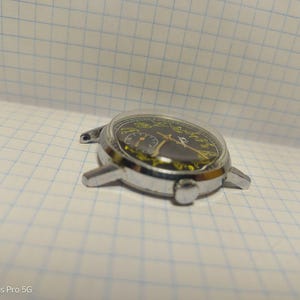 May include: A silver-toned wristwatch with a black face and yellow accents. The watch has a clear, domed crystal and a metal case with lugs for a strap. The watch is lying on a white grid paper.