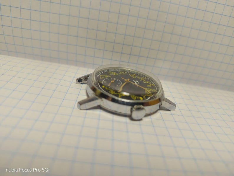 May include: A silver-toned wristwatch with a black face and yellow accents. The watch has a clear, domed crystal and a metal case with lugs for a strap. The watch is lying on a white grid paper.