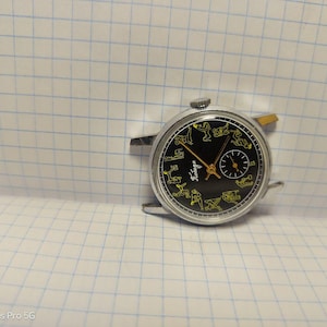 May include: Vintage wristwatch with a black face featuring yellow figures and hands. The watch has a silver-colored case and a small subdial. The word "Taviga" is printed on the watch face. The watch is set against a grid background.