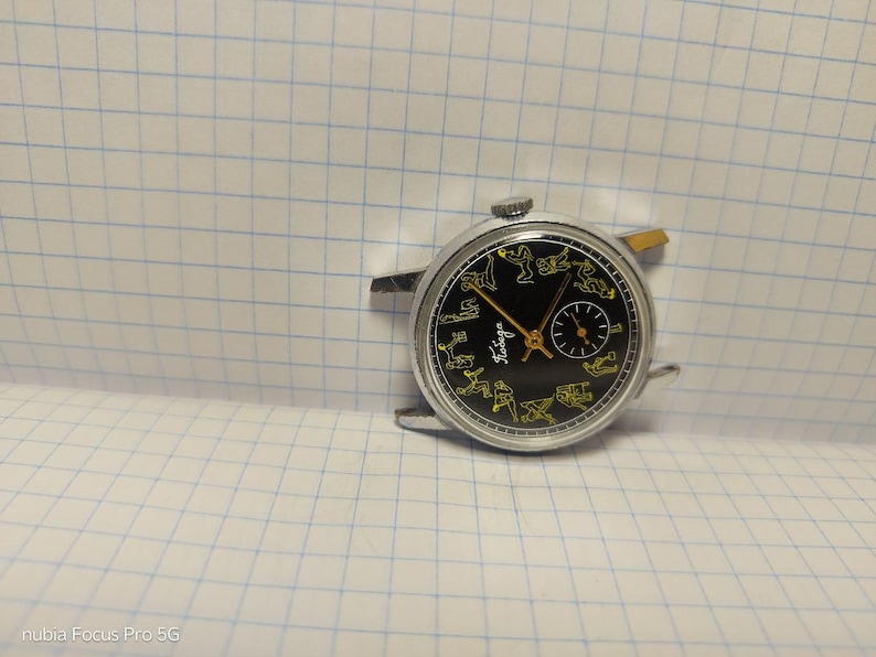 May include: Vintage wristwatch with a black face featuring yellow figures and hands. The watch has a silver-colored case and a small subdial. The word "Taviga" is printed on the watch face. The watch is set against a grid background.