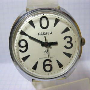 Puede incluir: Un reloj de pulsera plateado con una esfera blanca y números negros. El reloj tiene una correa de cuero negro y dice "Raketa" y "Сделано в СССР" en la esfera.