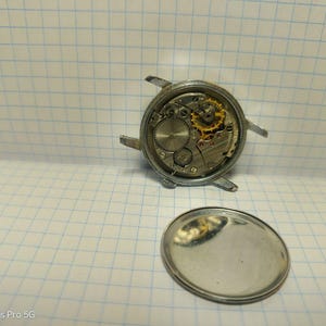 May include: A disassembled wristwatch showing the intricate internal mechanisms. The watch's silver casing is visible, along with the gears, springs, and other components. The back cover of the watch is also present, lying separately.