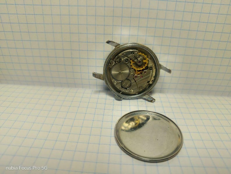 May include: A disassembled wristwatch showing the intricate internal mechanisms. The watch's silver casing is visible, along with the gears, springs, and other components. The back cover of the watch is also present, lying separately.