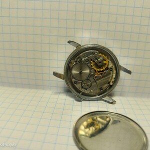 May include: Close-up of a disassembled mechanical watch, showcasing the intricate internal gears and components. The watch's silver casing is visible, along with the exposed movement, including gold-colored gears. The watch's back cover is also present.