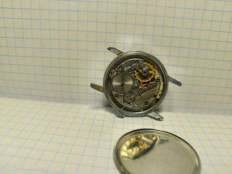 May include: Close-up of a disassembled mechanical watch, showcasing the intricate internal gears and components. The watch's silver casing is visible, along with the exposed movement, including gold-colored gears. The watch's back cover is also present.