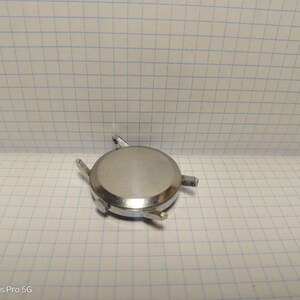May include: A silver-colored watch case with four lugs for attaching a strap. The round case has a brushed finish and is lying on a grid-patterned surface. The watch case appears to be a component for watch repair or restoration.