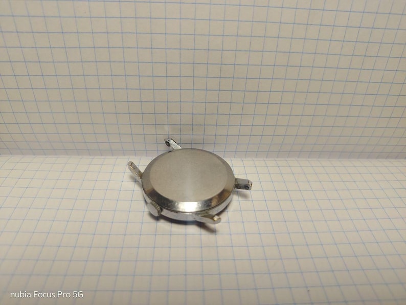 May include: A silver-colored watch case with four lugs for attaching a strap. The round case has a brushed finish and is lying on a grid-patterned surface. The watch case appears to be a component for watch repair or restoration.