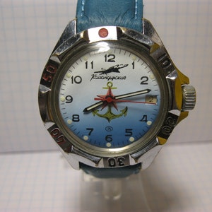 Puede incluir: Un reloj de pulsera plateado con una esfera azul y una aguja de segundos roja. La esfera del reloj tiene un ancla blanca y el texto "Командирские" en negro. El reloj tiene una correa de cuero azul.