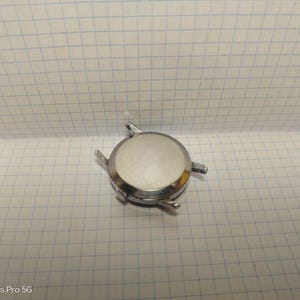 May include: A silver-colored watch case with four lugs for attaching a watch band. The case has a circular shape with a flat, reflective surface. The watch case is on a white grid paper.