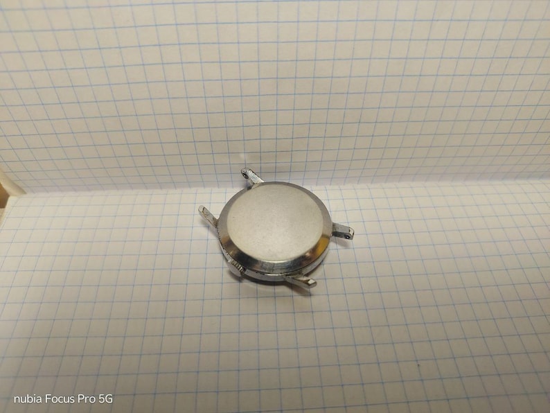 May include: A silver-colored watch case with four lugs for attaching a watch band. The case has a circular shape with a flat, reflective surface. The watch case is on a white grid paper.