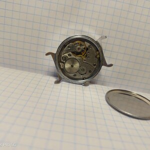 May include: A disassembled vintage watch with exposed gears and mechanisms. The watch's silver casing is open, revealing intricate internal components. A detached silver back cover lies nearby, showcasing the watch's internal workings.