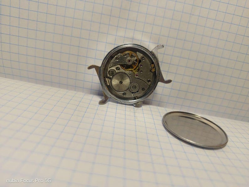 May include: A disassembled vintage watch with exposed gears and mechanisms. The watch's silver casing is open, revealing intricate internal components. A detached silver back cover lies nearby, showcasing the watch's internal workings.