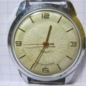 Puede incluir: Un reloj de pulsera plateado con una esfera blanca y agujas negras. El reloj tiene un diseño circular con marcadores de hora en números romanos y una ventana de fecha en la posición de las 3 en punto. La esfera del reloj también presenta el texto "СССР" y "МОСКВА".