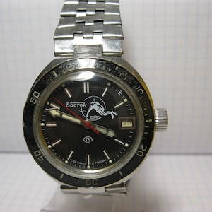 May include: A vintage Vostok wristwatch with a black dial and a stainless steel band. The watch face features white hands, a red second hand, and a diver graphic. The bezel has minute markers. The date is displayed at the 3 o'clock position.