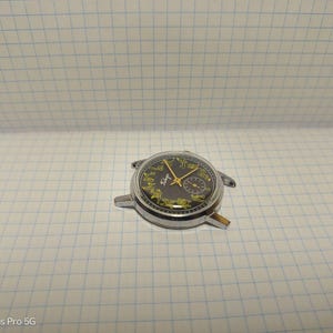 May include: A vintage wristwatch with a black face and gold hands. The watch has a silver-colored metal casing and a small subdial. The watch is missing its band and is resting on a white grid paper.