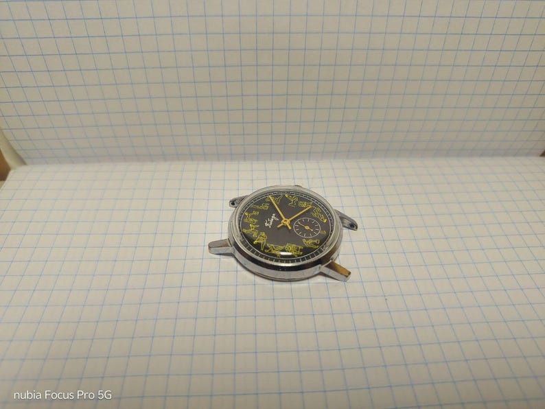 May include: A vintage wristwatch with a black face and gold hands. The watch has a silver-colored metal casing and a small subdial. The watch is missing its band and is resting on a white grid paper.