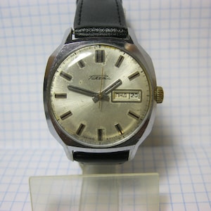 Puede incluir: Un reloj de pulsera plateado con una correa de cuero negro. La esfera del reloj es blanca con marcas negras y una ventana de fecha que muestra "ПНД 20".
