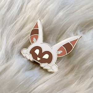 May include: A white and brown enamel pin of a cartoon animal with large ears peeking out from behind a surface. The animal has a brown face with white eyes and a small mouth.