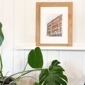 May include: A framed watercolour painting of a brick building with windows and a sign. The painting is on a white shelf with a green plant on either side.