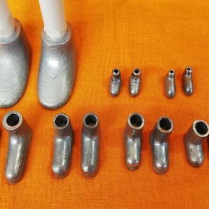 Op de afbeelding: Een set van 12 zilveren metalen schoenvormen, waaronder twee grote vormen en tien kleinere vormen. De vormen zijn allemaal van verschillende grootte en vorm en zijn in een rij op een fel oranje oppervlak gerangschikt.