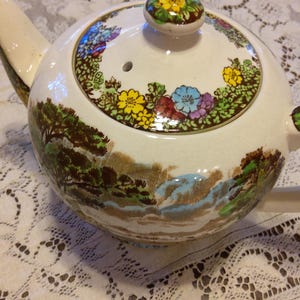 May include: A white ceramic teapot with a landscape design. The teapot features a lid with a floral pattern and a knob. The body of the teapot has a landscape scene with trees, water, and mountains.