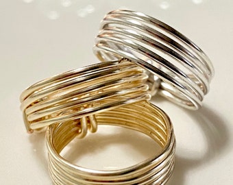 May include: Close-up of three wire-wrapped rings. One ring is silver, and two are gold. Each ring is made of multiple strands of wire, creating a textured, layered effect. The rings are displayed on a white surface, with a blurred background of white pearls.