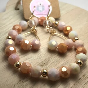 May include: A pair of gold hoop earrings with peach, white, and iridescent glass beads. The earrings are made with a gold wire and have a dangling charm.