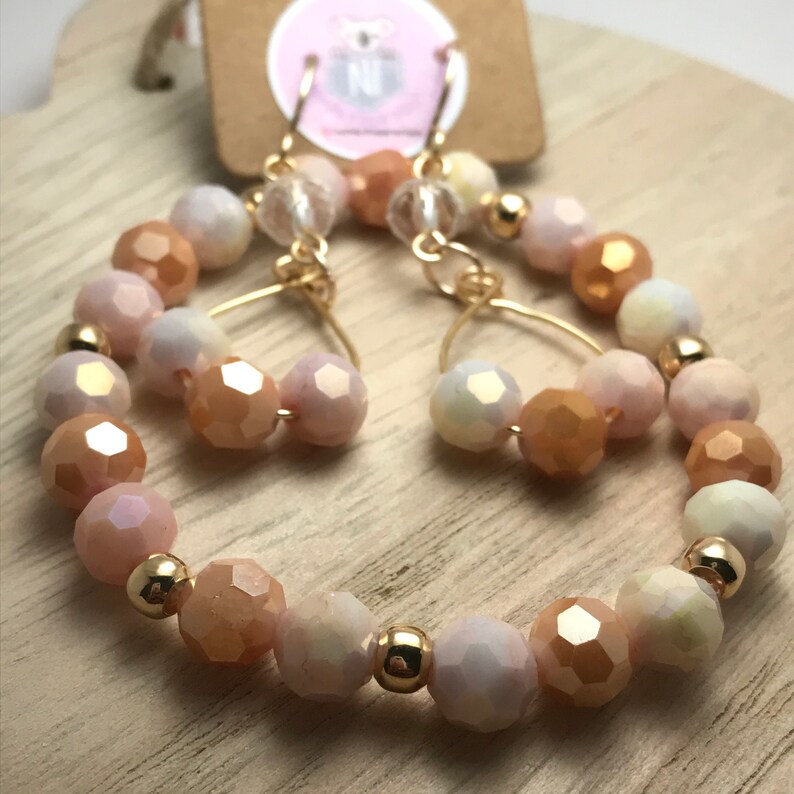 May include: A pair of gold hoop earrings with peach, white, and iridescent glass beads. The earrings are made with a gold wire and have a dangling charm.