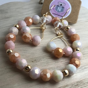 May include: A pair of gold hoop earrings with peach, white, and iridescent glass beads. The earrings are made with a wire hoop and have a dangling charm.