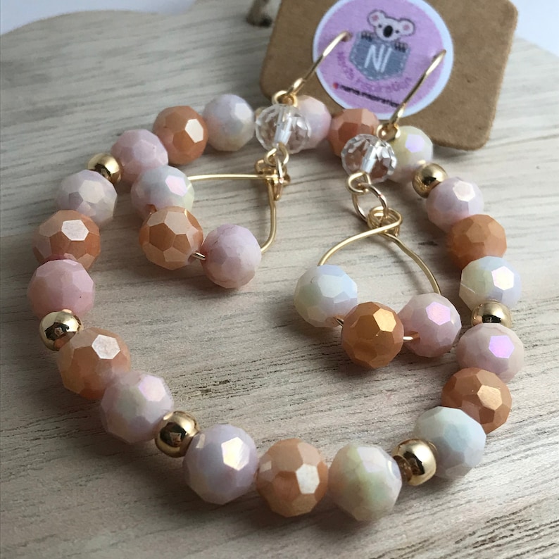 May include: A pair of gold hoop earrings with peach, white, and iridescent glass beads. The earrings are made with a wire hoop and have a dangling charm.