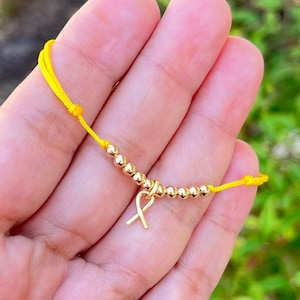 Op de afbeelding: Een gele armband van touw met gouden kralen en een gouden lintbedel.