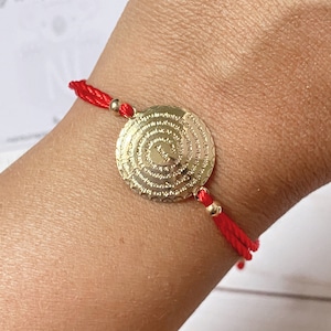 May include: A red string bracelet with a gold circular charm featuring a prayer inscribed in a circular pattern. The charm has a raised center with the word "Amen" inscribed.