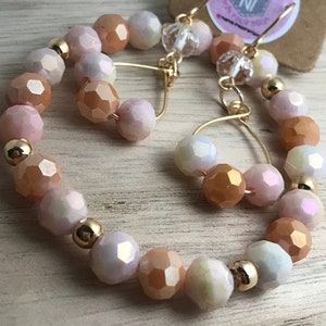 May include: A pair of gold hoop earrings with iridescent peach and white beads. The earrings are made with a wire hoop and have a delicate, feminine look.