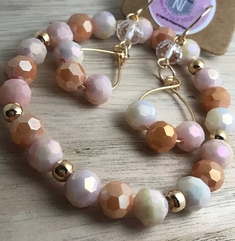 May include: A pair of gold hoop earrings with iridescent peach and white beads. The earrings are made with a wire hoop and have a delicate, feminine look.