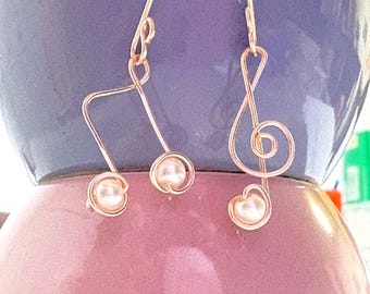 Op de afbeelding: Een paar oorbellen van roségoudkleurig draad met muzikale symbolen. De ene oorbel heeft de vorm van een muzieknoot en de andere van een vioolsleutel. Elke oorbel is versierd met kleine, ronde, witte kunstparels verwerkt in het draadontwerp.