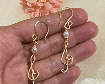Op de afbeelding: Een paar gouden draad oorbellen met een treble clef ontwerp en een witte parel accent.