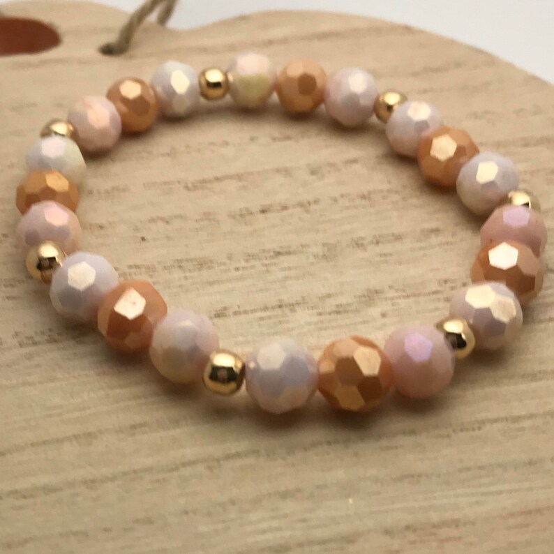 May include: A delicate bracelet made with iridescent white and peach-colored faceted beads and gold-toned spacers.