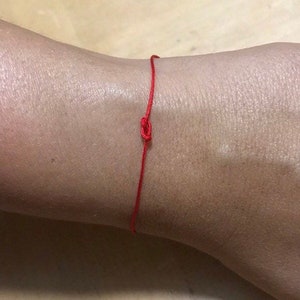 May include: A red string bracelet with a simple knot. The bracelet is tied around a person's wrist.