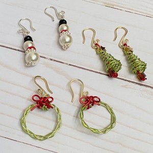 May include: Four pairs of Christmas-themed earrings. Two pairs feature a snowman and a Christmas tree design with white and green beads. The other two pairs feature a green wreath with a red bow.