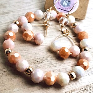 May include: A pair of gold hoop earrings with peach and white iridescent glass beads. The earrings are made with a wire hoop and have a dangling charm.