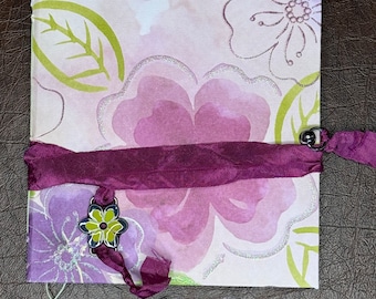 May include: A small, white journal with a floral design and a purple ribbon tied around it. The journal has a green leaf design and a purple flower design. A small green and yellow flower charm is attached to the ribbon.