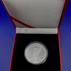 May include: A silver American Eagle coin in a red velvet box with a black velvet interior. The coin features a profile of Lady Liberty with the inscription "Liberty" and "In God We Trust".