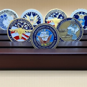 May include: A wooden coin display stand with six slots, holding six different military challenge coins. The coins feature various designs, including an eagle, a fighter jet, and a cartoon sailor.