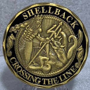 May include: A gold-colored coin with a black background. The coin features a depiction of a mythical sea creature with a trident and the words "Shellback Crossing the Line".