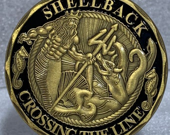 May include: A gold-colored coin with a black background. The coin features a depiction of a mythical sea creature with a trident and the words "Shellback Crossing the Line".